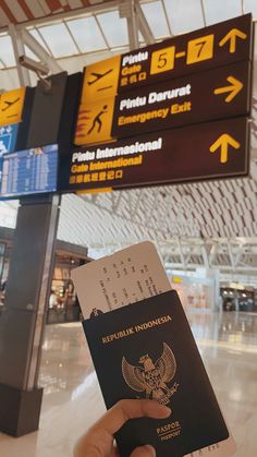 a person holding up a passport in front of an airport sign with directions to different destinations