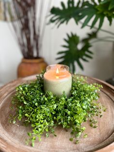 a lit candle sitting on top of a wooden tray filled with green plants and greenery