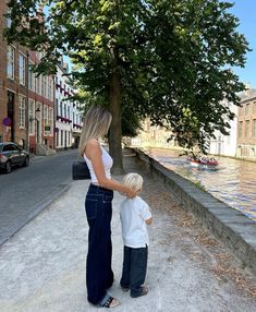 a woman standing next to a little boy in front of a tree on the side of a river