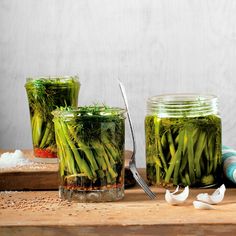 three mason jars filled with green vegetables on top of a wooden cutting board next to garlic