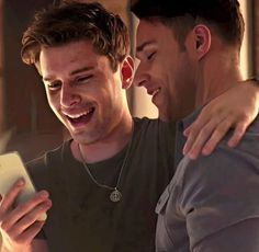 two young men laughing and looking at their cell phones while one holds his arm around the other's shoulder