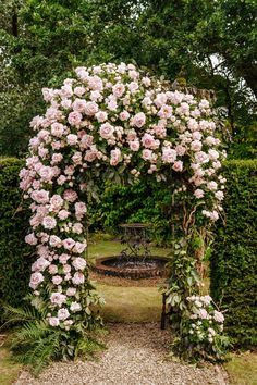 an arch covered with pink flowers in the middle of a gravel path surrounded by hedges