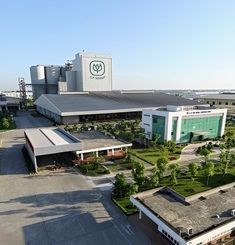 an aerial view of a factory building with trees and buildings in the foreground, on a sunny day