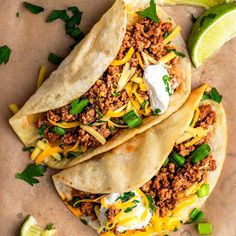 three tacos with ground beef, cheese and sour cream on them next to lime wedges