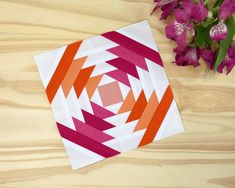 an orange, pink and white quilt next to purple flowers on a wooden table top