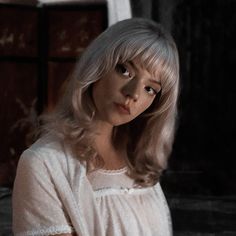 a woman with blonde hair wearing a white dress and looking at the camera while standing in front of a dresser