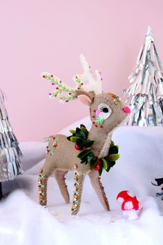 a stuffed reindeer is standing in the snow next to a christmas tree and mushroom house