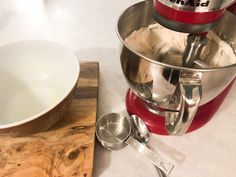 a red mixer sitting on top of a wooden cutting board next to a white bowl