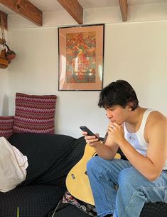 a young man sitting on top of a couch holding a guitar and looking at his cell phone