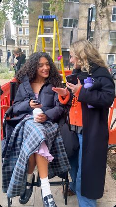 two women sitting in a wheel chair on the sidewalk talking to each other and one holding a cup