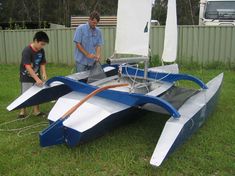 two men working on a small sailboat in the grass with another man standing next to it