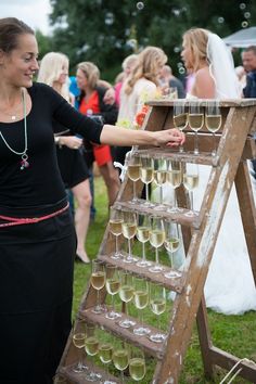 a woman standing next to a wooden ladder filled with wine glasses on top of a lush green field