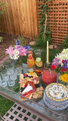 a table with plates and glasses on it in front of a wooden fence, surrounded by flowers