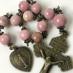 Handmade traditional Catholic single decade tenner rosary using pink petrified rhodonite smooth round stone beads. Antique bronze daisy bead caps on every bead with additional flower bead caps on Our Father bead. Antique bronze heart shaped Miraculous Medal center with Pardon Crucifix. Hail Mary beads are 8mm with the Our Father bead being 10mm. Designed to be used and/or displayed. Measures about 10.5" long. This rosary would make a beautiful gift for a baby shower, Baptism, First Communion, Confirmation, Valentine's Day, Anniversary or any other special event or occasion. Comes in a beautiful black velvet pouch. I ship same or next business day. All of my rosaries are one of a kind and are my own personal creation. Photos are a part of the description-you will receive the exact rosary pi Affordable Pink Rosary Gift, Spiritual Pink Spacer Beads, Pink Natural Stones Beads As Gift, Pink 8mm Bohemian Beads, Vintage Rosary With Round Beads As Gift, Spiritual Rosary Bracelet With Polished Beads, Pink Bohemian Rosary Bracelet Gift, Bohemian Rosary With Polished Beads As Gift, Rosary With Natural Stones For Meditation