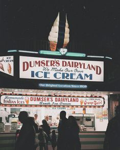 some people are standing in front of a ice cream shop at night with the lights on