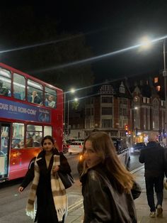 two women walking down the street in front of a red double decker bus at night