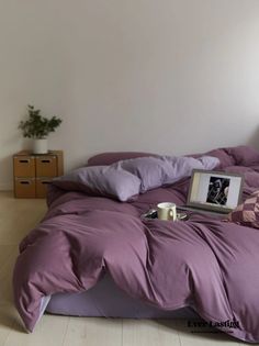 a bed with purple sheets and pillows in a room next to a plant on the floor