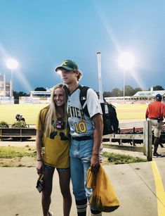 a man and woman standing next to each other on a baseball field with lights in the background