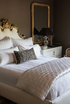 a bed with white sheets, pillows and a gold framed mirror on the wall above it