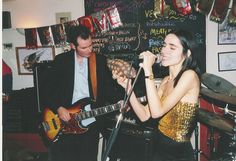 a woman singing into a microphone while holding a guitar in front of a man playing the guitar