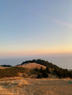 the sun is setting over some hills with trees on top and an ocean in the distance