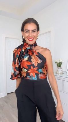 a woman standing in front of a white wall wearing black pants and a floral top