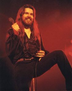 a man with long hair is sitting on a stool and holding a microphone in his hand