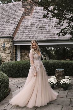 a woman standing in front of a house wearing a wedding dress with long sleeves and flowers