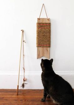 a black cat sitting on the floor looking at a wall hanging with a wooden stick