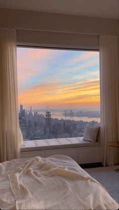 a bed sitting next to a window in a bedroom under a sunset sky line with the city skyline