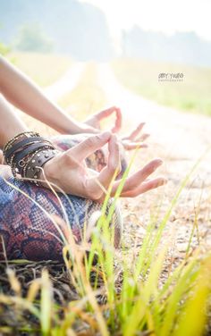 a woman sitting on the ground doing yoga in front of grass and dirt road with her hands up