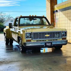 an old yellow truck parked in front of a building on a wet parking lot next to a brick wall