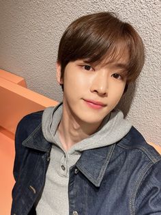 a young man wearing a denim jacket sitting on a wooden bench in front of a wall