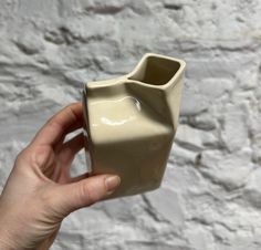 a hand holding a white ceramic vase in front of a stone wall with no one around it