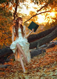 a woman in a white dress is sitting on a fallen tree and holding a book