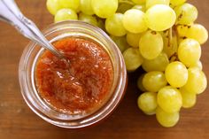 a glass jar filled with jam next to grapes on a wooden table and a spoon