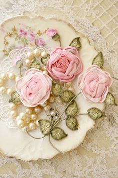 three pink roses are on a plate with pearls and lace around the edges, sitting on a tablecloth