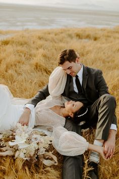 a bride and groom sitting on the ground in tall grass by the water at their wedding