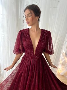 This stunning burgundy gown features a captivating plunging V-neckline, delicately pleated bodice, and romantic puffed sleeves. The shimmering lace fabric drapes gracefully into a flowing A-line silhouette, cinched at the waist for a flattering fit. Perfect for evening galas or any special occasion, this dress combines elegance with a touch of enchantment, making it an unforgettable choice. Lace Evening Dresses Short, Burgundy Floor-length Gala Gown, Burgundy Floor-length Ball Gown For Wedding, Floor-length Burgundy Dress With Fitted Bodice, Luxury Burgundy A-line Dress, Burgundy Corset, Burgundy Floor-length Evening Gown, Teuta Matoshi Dresses, Matoshi Dress