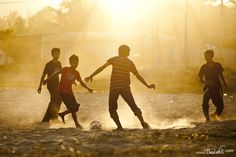 three young boys playing soccer on the beach at sunset, with sun shining behind them