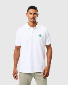 A man stands against a plain background, wearing the Psycho Bunny MENS COURTSIDE PIQUE POLO - B6K655C200. This tennis-inspired white embroidered pique polo features a small green logo on the left chest and is crafted from soft Pima cotton. It has a collar and short sleeves, which he pairs with light-colored pants. The man has short, dark hair and maintains a neutral expression. Classic White Polo Shirt With Embroidered Logo, White Sporty Polo Shirt With Embroidered Logo, White Cotton Polo Shirt With Polo Collar, Sporty Polo Shirt With Embroidered Logo, Sporty Collared Polo Shirt With Embroidered Logo, White Preppy Polo Shirt, White Cotton Preppy Polo Shirt, Preppy White Cotton Polo Shirt, White Cotton Collared Polo Shirt