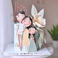 a birthday cake decorated with two people and flowers on top of a white shelf next to a potted plant