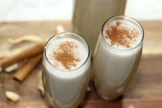 two glasses filled with milk and cinnamon on top of a wooden table next to bottles