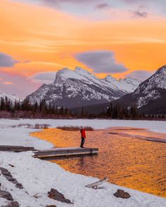 a person standing on a dock in the snow near some water and mountains at sunset