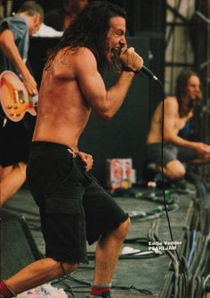 a shirtless man with long hair and no shirt on singing into a microphone while holding a guitar in his right hand