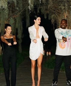 two women and a man standing on a walkway in front of trees with lights behind them