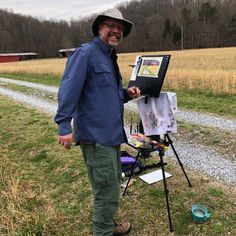 a man standing next to an easel in a field with a painting on it