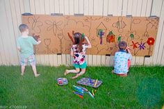 three children are playing in the grass near a wall with drawings on it and crayons