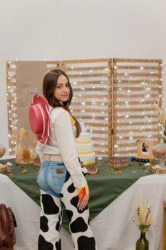 a woman standing in front of a table wearing cow print pants and a cowboy hat