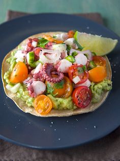 an open pita topped with vegetables and meat on top of a blue plate next to a lemon wedge